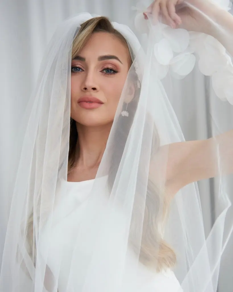 Luxuriöses Portrait einer blonden Braut aus einem Hochzeits-Styling-Workshop von Alexandra Lederer. Sie blickt durch einen feinen Tüllschleier, während die floralen Valeria Ohrhänger von Sternenauge mit ihren handgeformten weißen Blüten perfekt zur Geltung kommen.