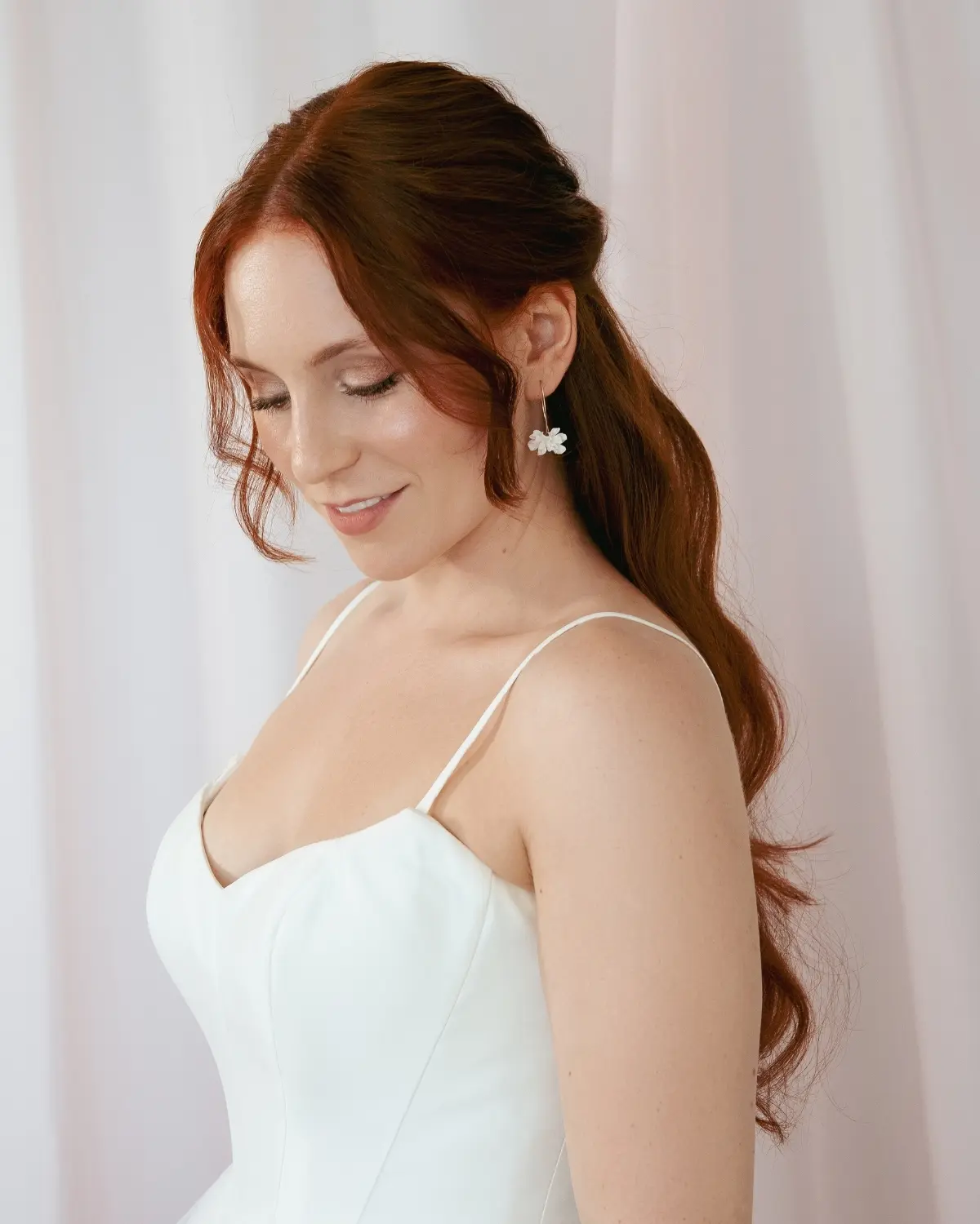 Eine lächelnde Braut mit roten, halb hochgesteckten Haaren im weißen Hochzeitskleid trägt die filigranen Valeria Ohrhänger von Sternenauge. Das Bild im hellen Boho-Stil zeigt den modernen Braut-Look von der Seite.
