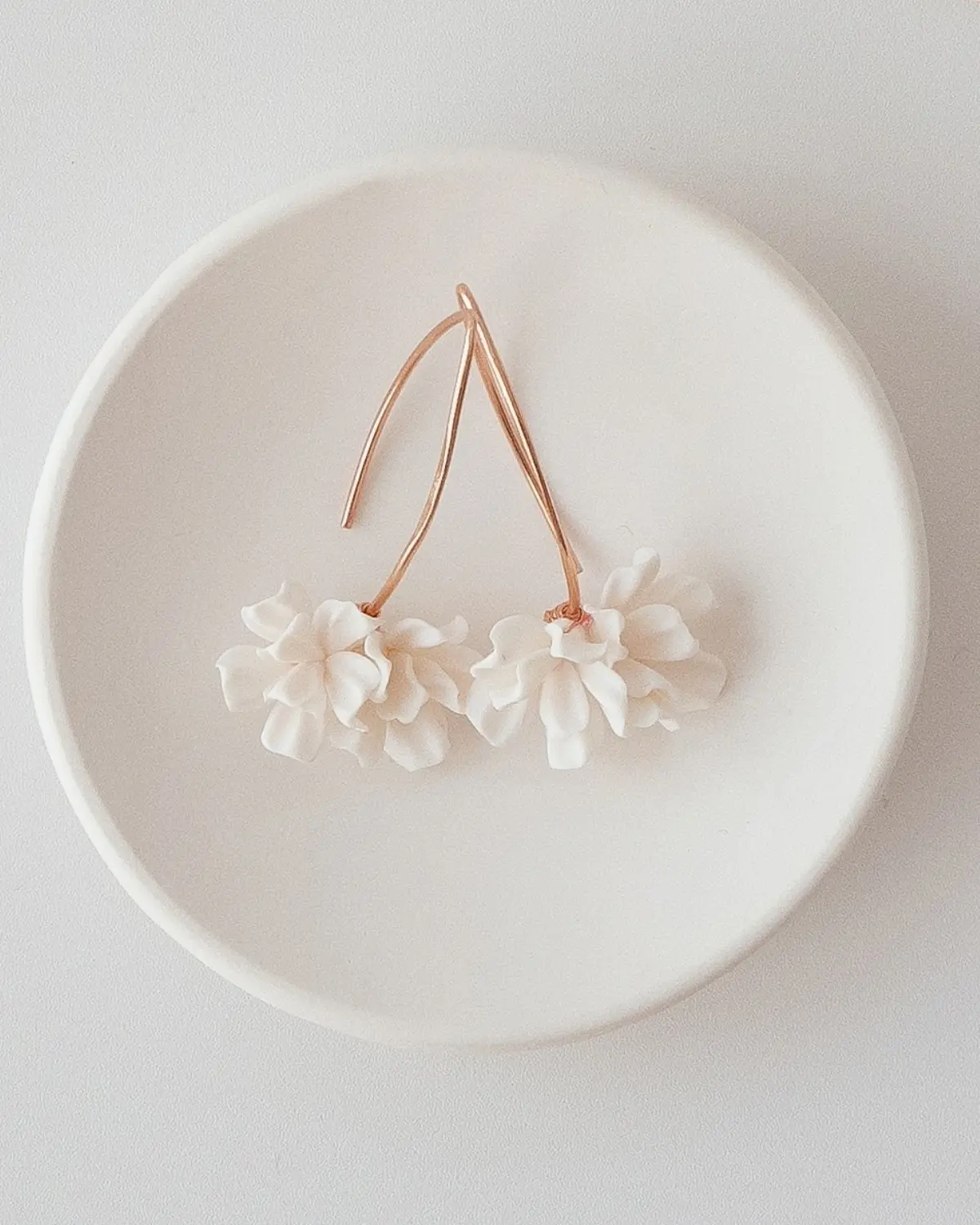 Die Valeria Ohrhänger aus 925 Sterling Silber mit 18K Roségoldvergoldung liegen in einer minimalistischen weißen Keramikschale. Gut zu sehen sind die handgeformten weißen Blüten aus Polymer Clay.