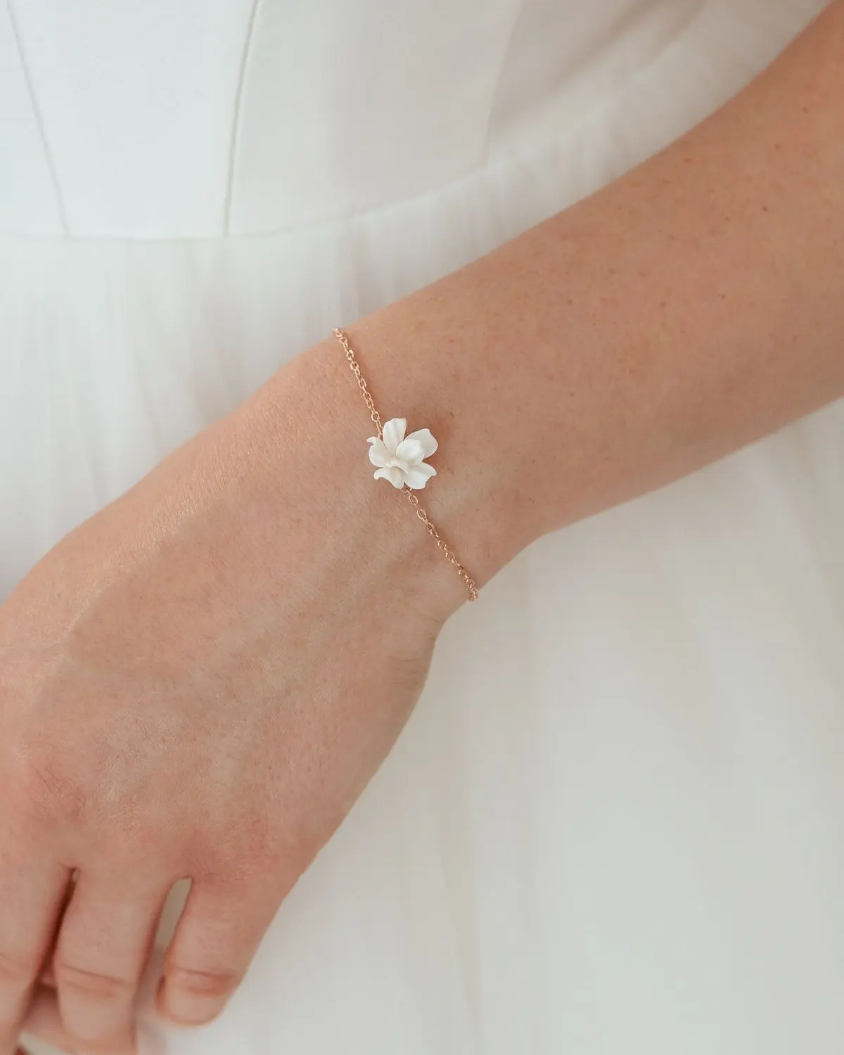 Zartes florales Sternenauge Valeria Armband in Roségold-Ton mit weißer Polymer-Clay-Blüte, getragen am Handgelenk einer Braut vor einem weißen Hochzeitskleid.