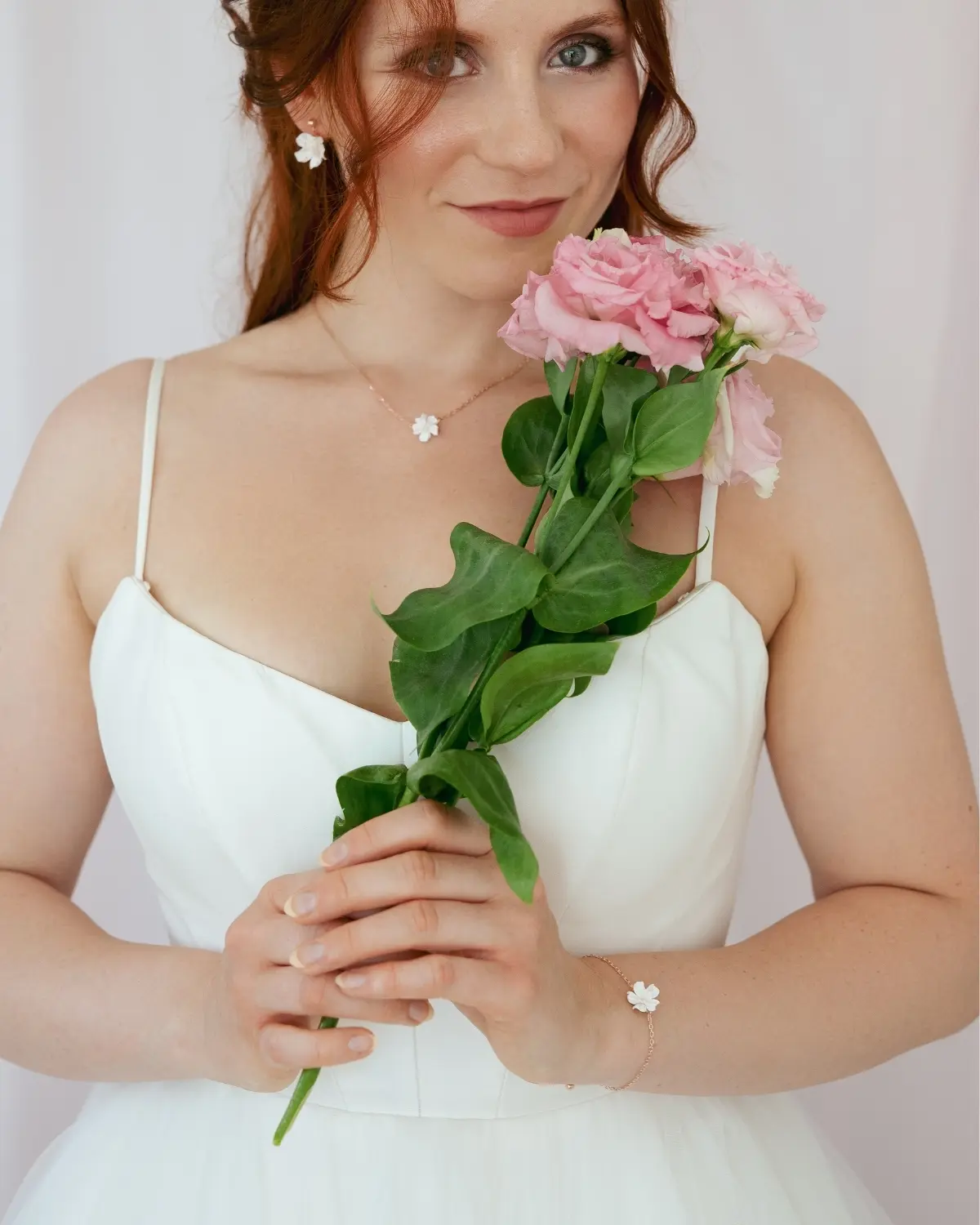 Eine Braut im weißen Hochzeitskleid hält rosa Blumen und zeigt den Schmuck-Look der Sternenauge Valeria Kollektion. Sie trägt die Halskette aus 18k rosévergoldetem 925 Sterling Silber kombiniert mit den passenden Ohrringen und dem Armband. Styling-Beispiel – Ohrringe und Armband separat erhältlich.