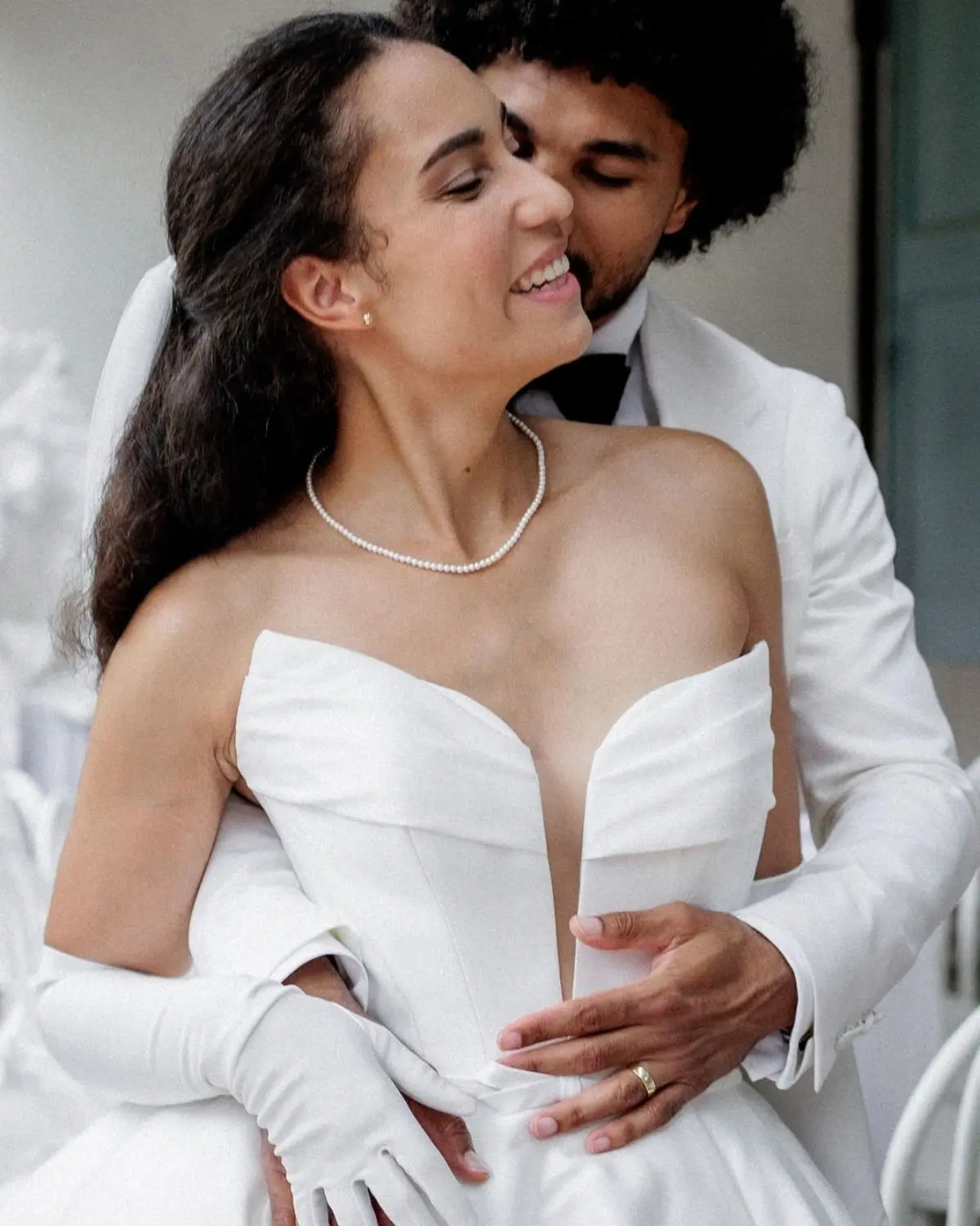 Glückliche Braut im modernen Hochzeitskleid mit der klassischen Annouk Perlenkette, umarmt von ihrem Bräutigam.