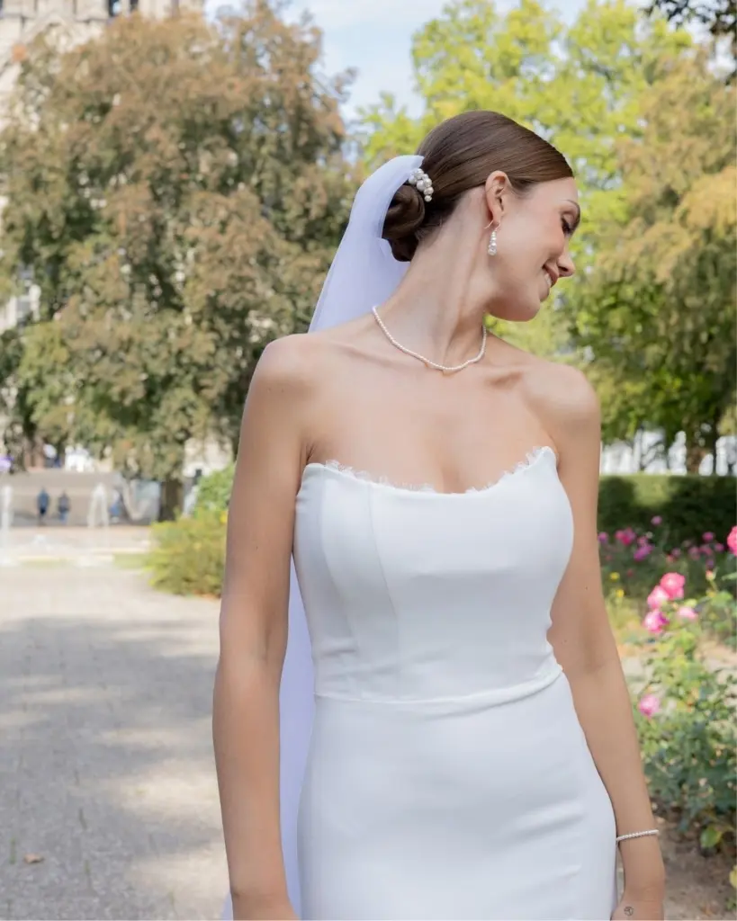 Inspirationsfoto einer lächelnden Braut mit Schleier und modernem, trägerlosen Satin-Brautkleid, die das komplett aufeinander abgestimmte Sternenauge Annouk Schmuckset aus Halskette, Full-Pearl-Armband, Perlen-Ohrhänger und Haarkamm trägt.
