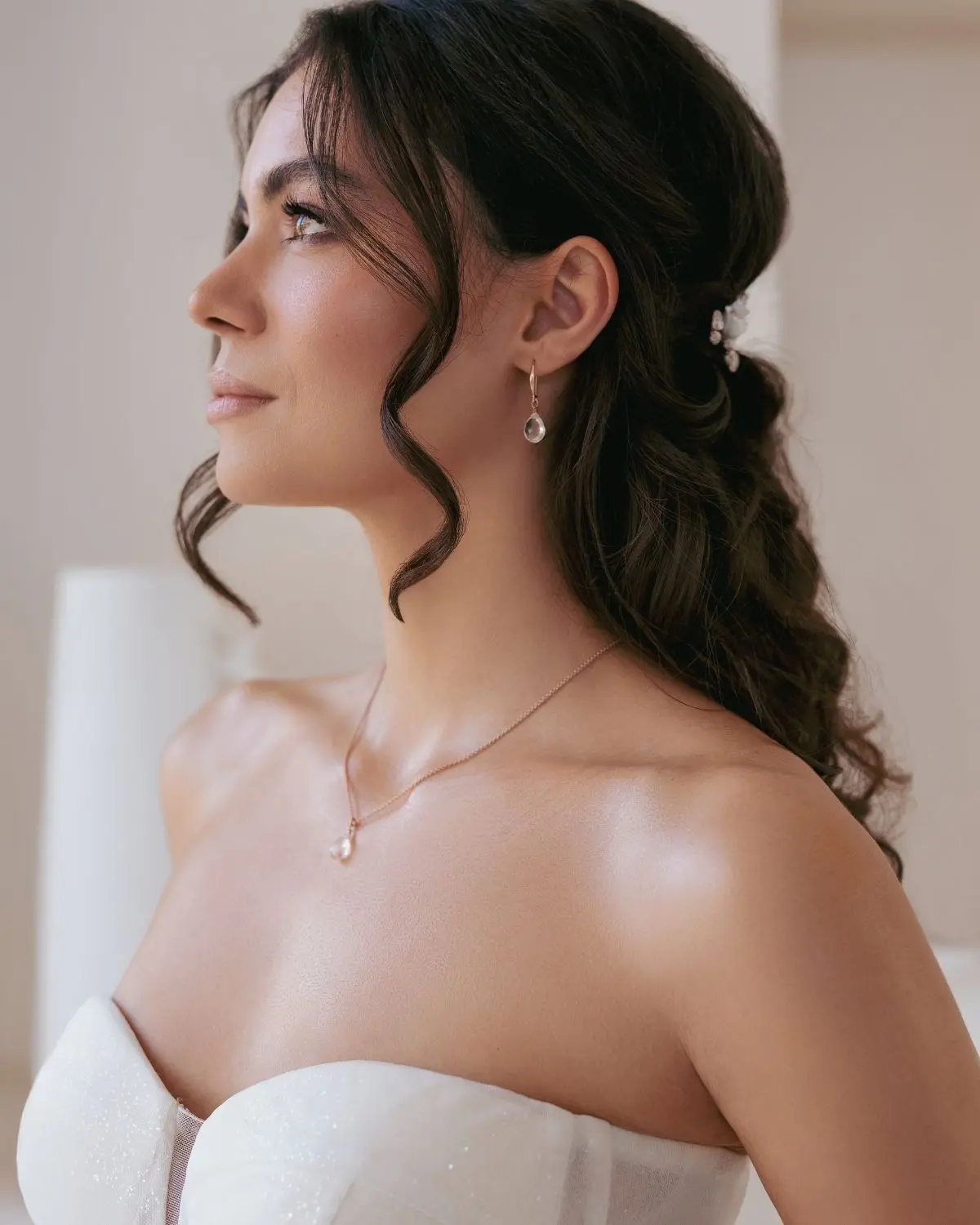 Seitenansicht einer Braut mit eleganter Hochzeitsfrisur und Haarschmuck, die das roségold-vergoldete Brautschmuck-Set Kyra von Sternenaufe mit Bergkristall-Halskette und tropfenförmigen Ohrhängern zum trägerlosen Hochzeitskleid trägt.