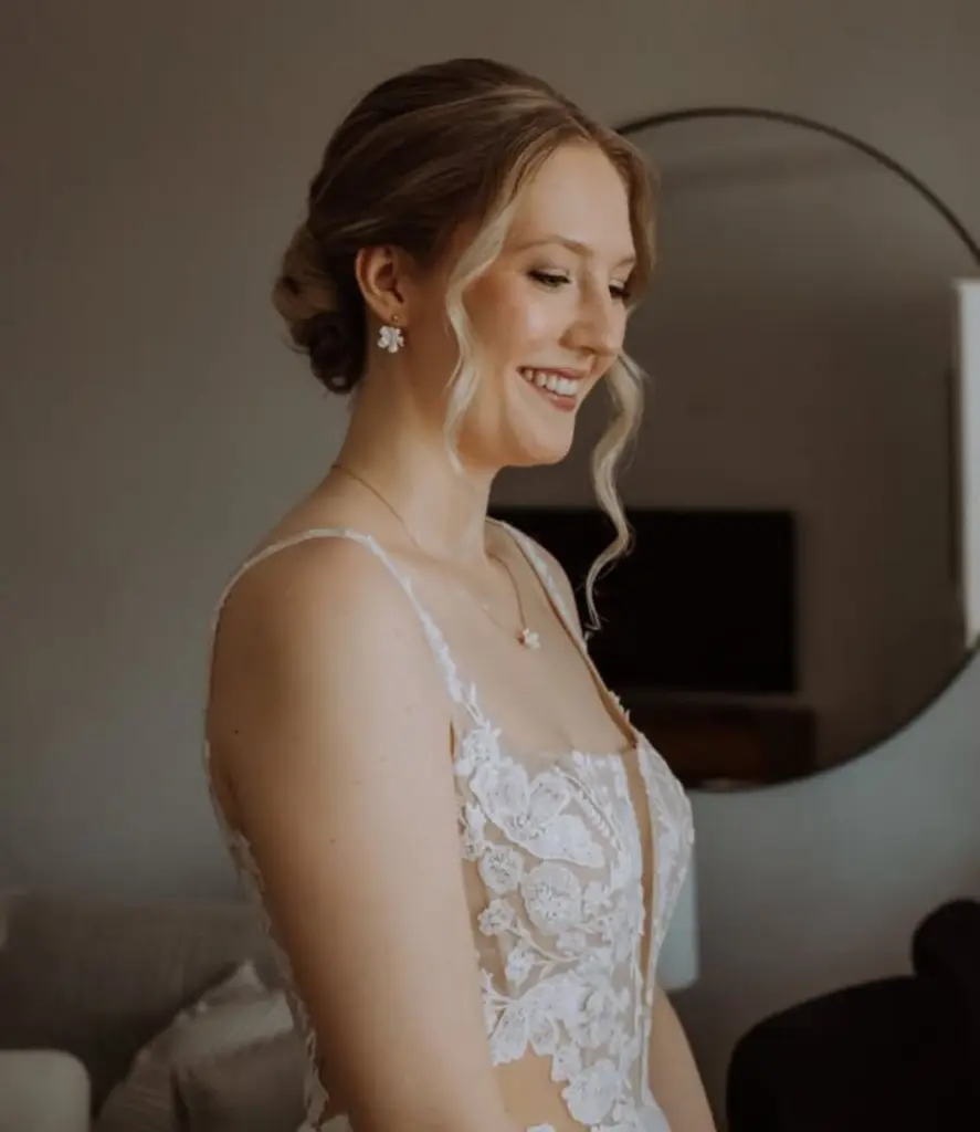 Seitliches Porträt einer lächelnden Braut mit eleganter Hochsteckfrisur und gelockten Strähnen. Sie trägt die filigrane Halskette und die passenden floralen Ohrstecker aus dem Valeria III Set zu einem modernen Brautkleid mit transparentem Ausschnitt und floralen Spitzen-Applikationen