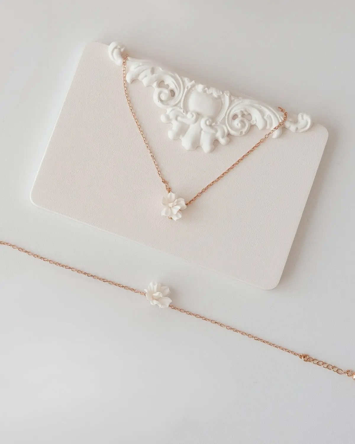 Präsentation der Valeria Halskette und des passenden Armbands aus dem Valeria Brautschmuck-Set III von Sternenauge auf einem hellen Hintergrund mit barockem Stuck-Zierelement. Die filigranen Ankerketten sind aus rosévergoldetem 925 Sterling Silber und mit jeweils einer dreidimensionalen, handgefertigten Blüte besetzt.