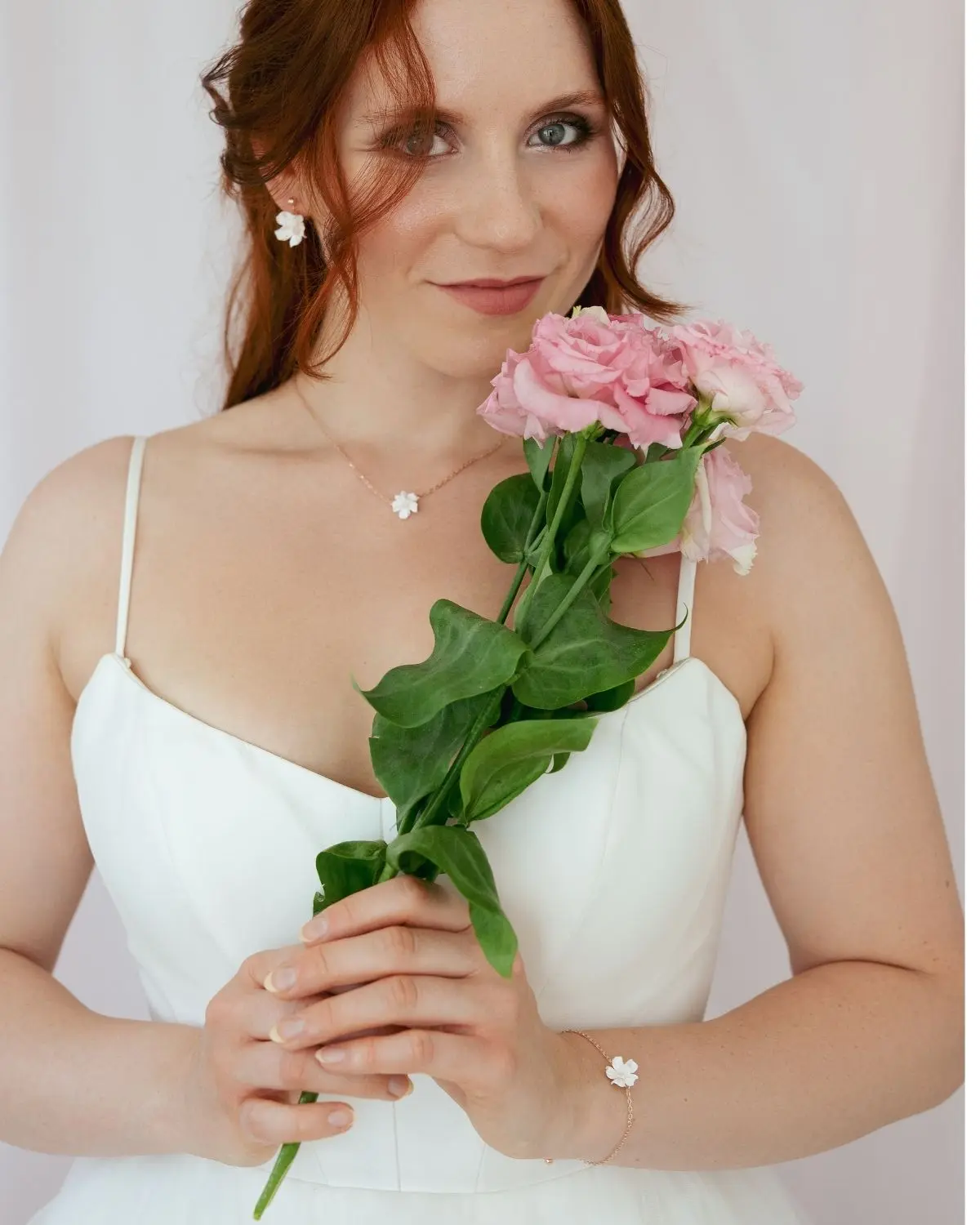 Porträt einer lächelnden Braut im weißen Hochzeitskleid mit Corsagen-Ausschnitt, die rosa Blumen hält. Sie trägt das komplette Valeria III Set bestehend aus floralen Ohrhängern, einer feinen Halskette und einem passenden Gliederarmband sowie Haarnadeln mit Blüten .