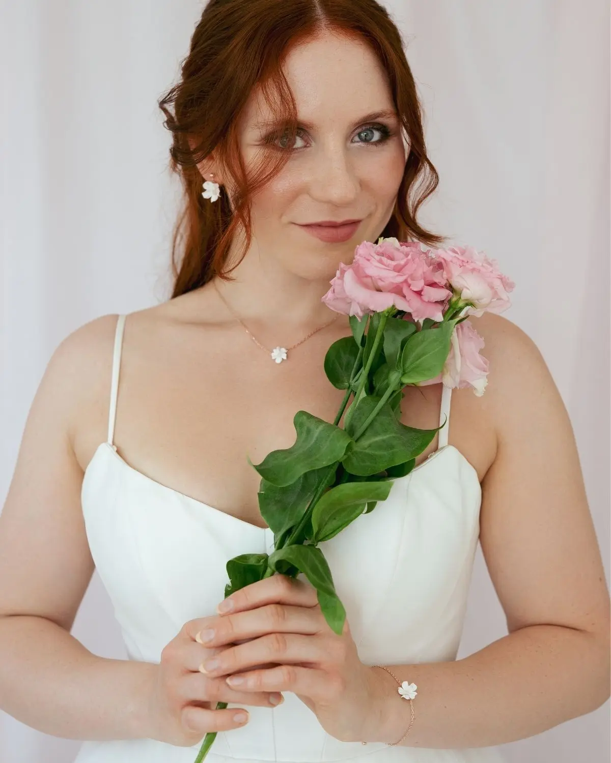 Eine rothaarige Braut im Hochzeitskleid mit einem rosa Blumenstrauß, die das dreiteilige Sternenauge Brautschmuck-Set Valeria mit Halskette, Ohrsteckern und Armband aus 925 Sterling Silber 18k rosévergoldet trägt.